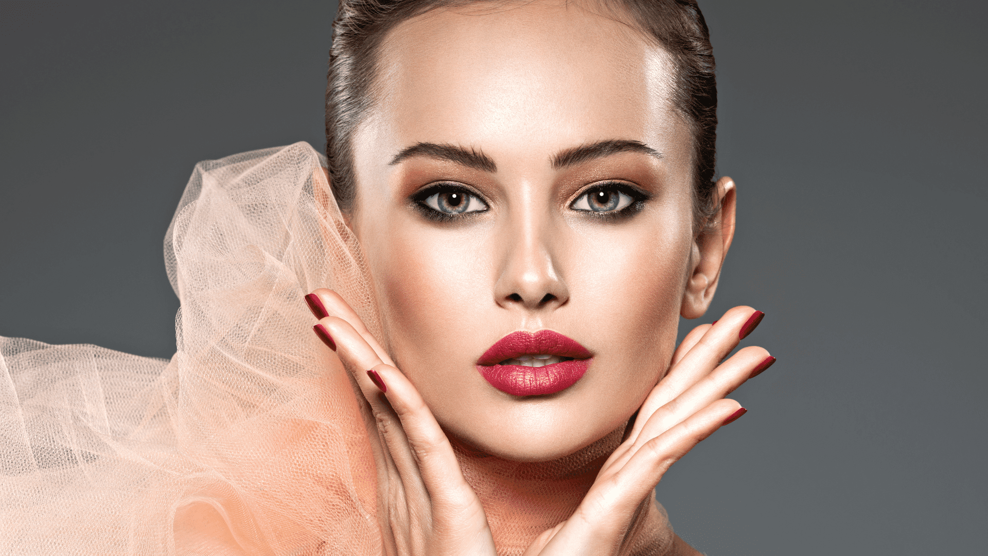 Close-up of a woman with elegant makeup, red lipstick, and nails, framed by peach tulle fabric.
