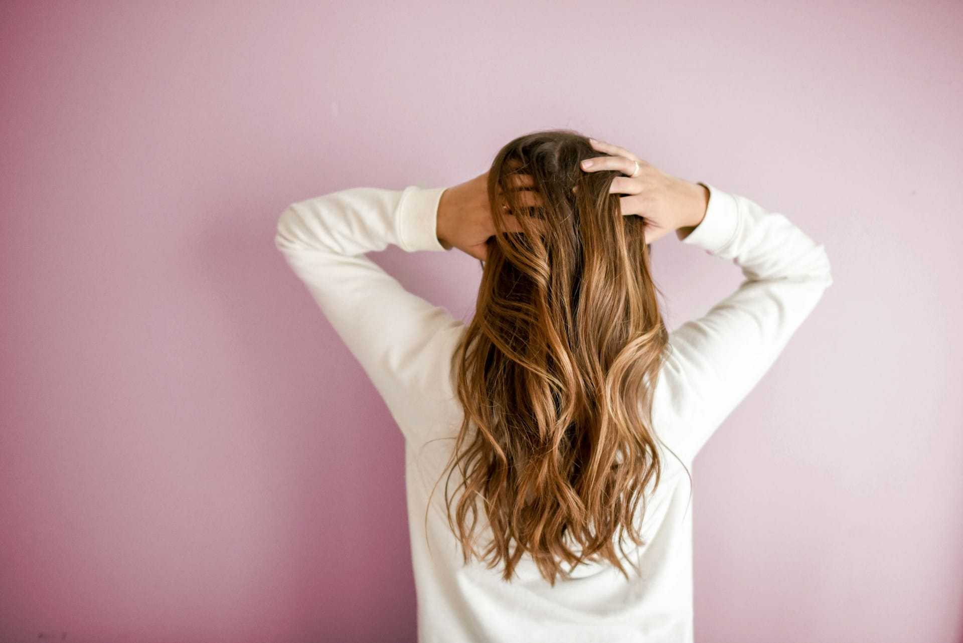 Person with long, wavy hair facing away and touching their head against a pink background.