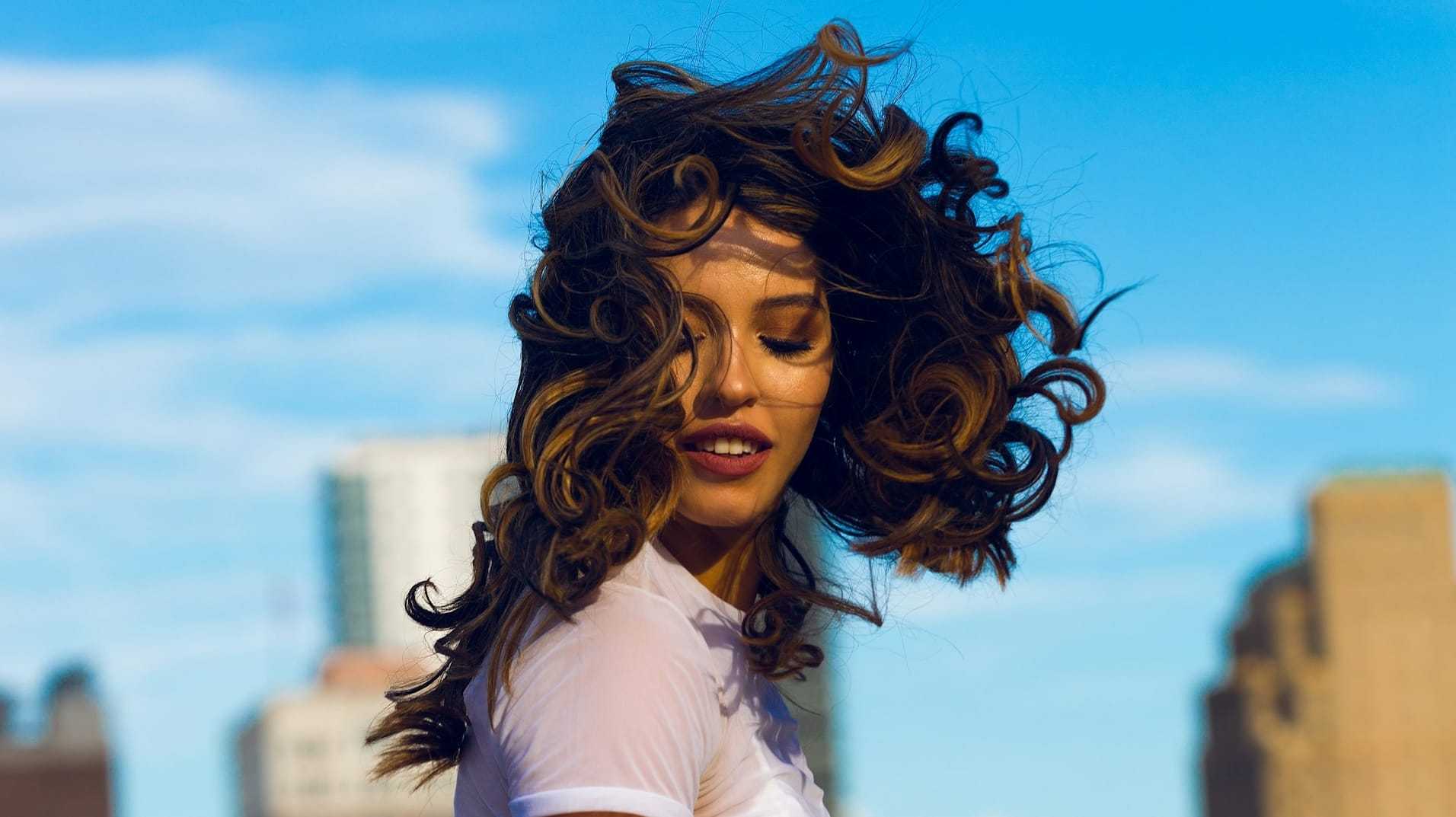 Woman with curly hair smiling against a cityscape and blue sky backdrop.